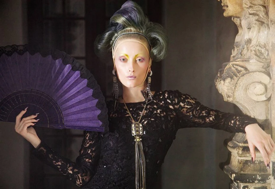 Elegant woman in black lace dress and elaborate makeup poses with a purple hand fan in an indoor setting with a classical architectural backdrop.