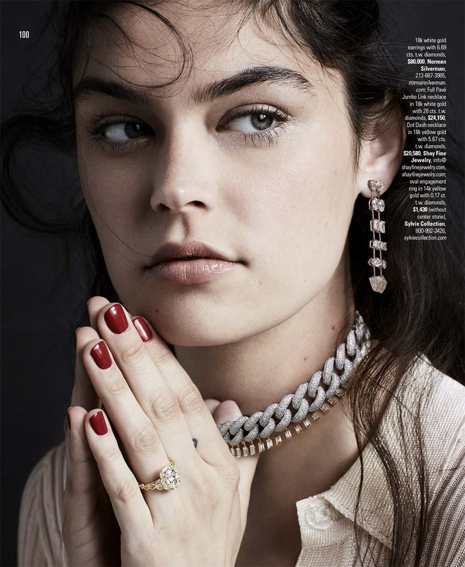 Close-up of a woman with dark hair, wearing a white top and diamond jewelry including a ring, necklace, and dangling earrings.