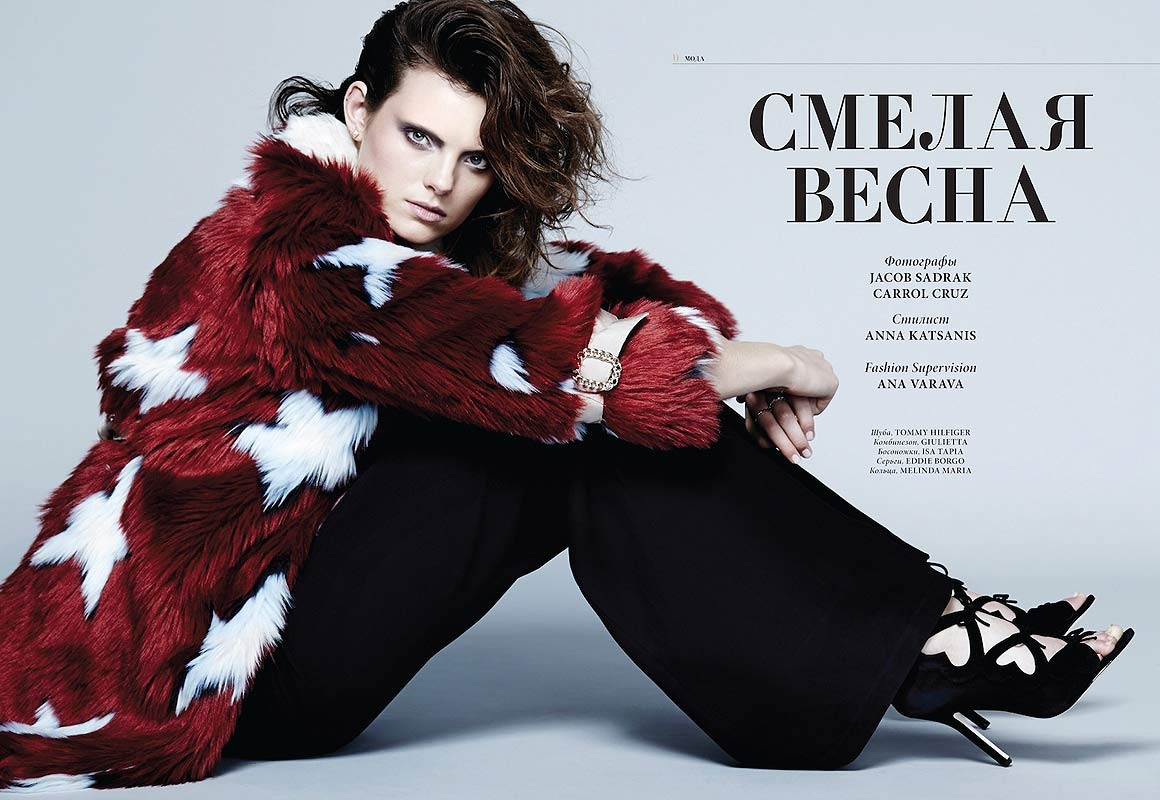 Model seated against a grey backdrop wearing a red and white patterned faux fur coat and black trousers