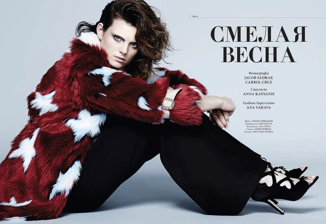 Model seated against a grey backdrop wearing a red and white patterned faux fur coat and black trousers