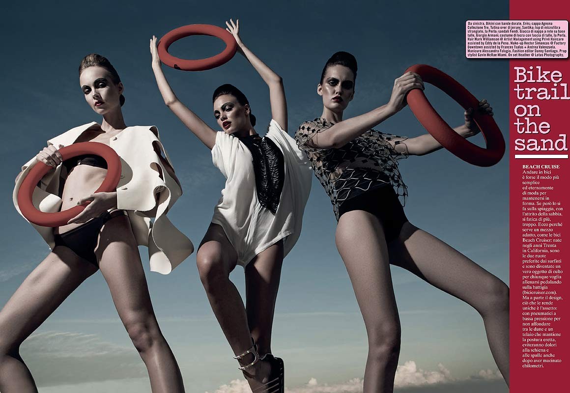 Three women in swimwear and outerwear hold red rings against a blue sky. Fashion editorial with modern poses and dramatic makeup.