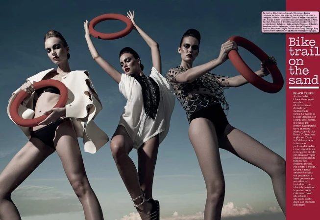 Three women in swimwear and outerwear hold red rings against a blue sky. Fashion editorial with modern poses and dramatic makeup.