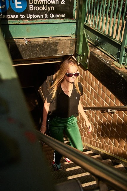 A woman in a black shirt and green pants walks up stairs from a subway station with a sign that says 'Downtown & Brooklyn'.