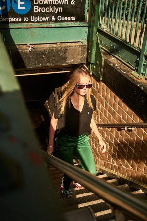A woman in a black shirt and green pants walks up stairs from a subway station with a sign that says 'Downtown & Brooklyn'.