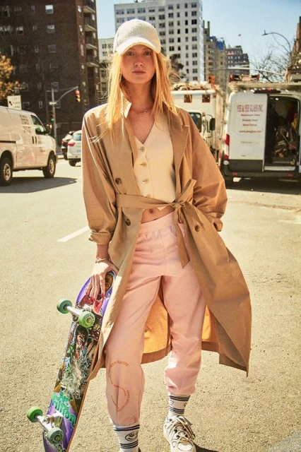 A young adult woman stands on a city street with skateboard, wearing beige coat, top, pink pants, sneakers and cap; daytime urban fashion.
