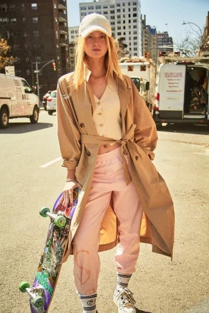 A young adult woman stands on a city street with skateboard, wearing beige coat, top, pink pants, sneakers and cap; daytime urban fashion.