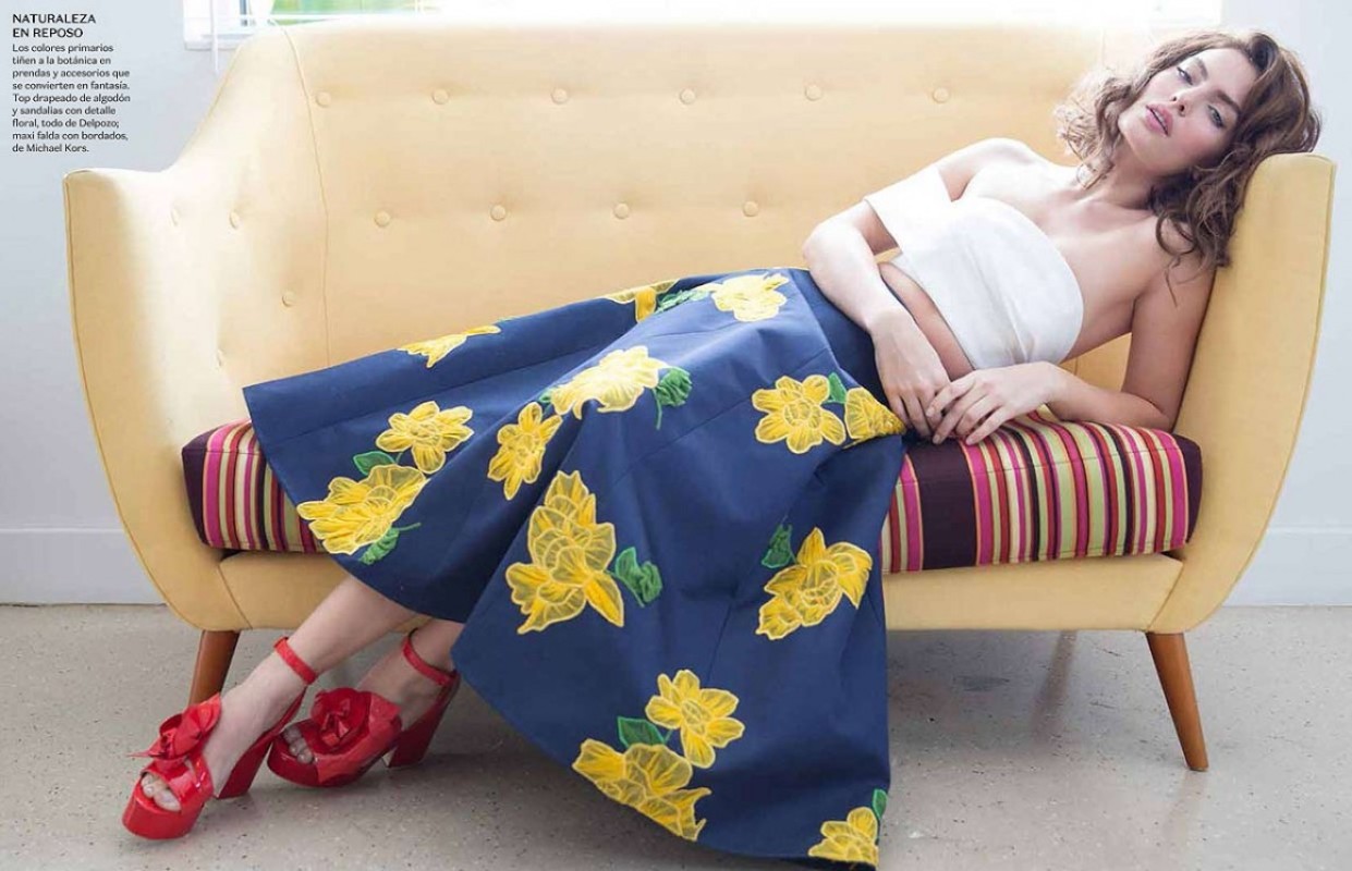 Woman in floral skirt and red shoes lounges on a yellow couch indoors; fashion editorial with bright, summery colors and retro vibes.