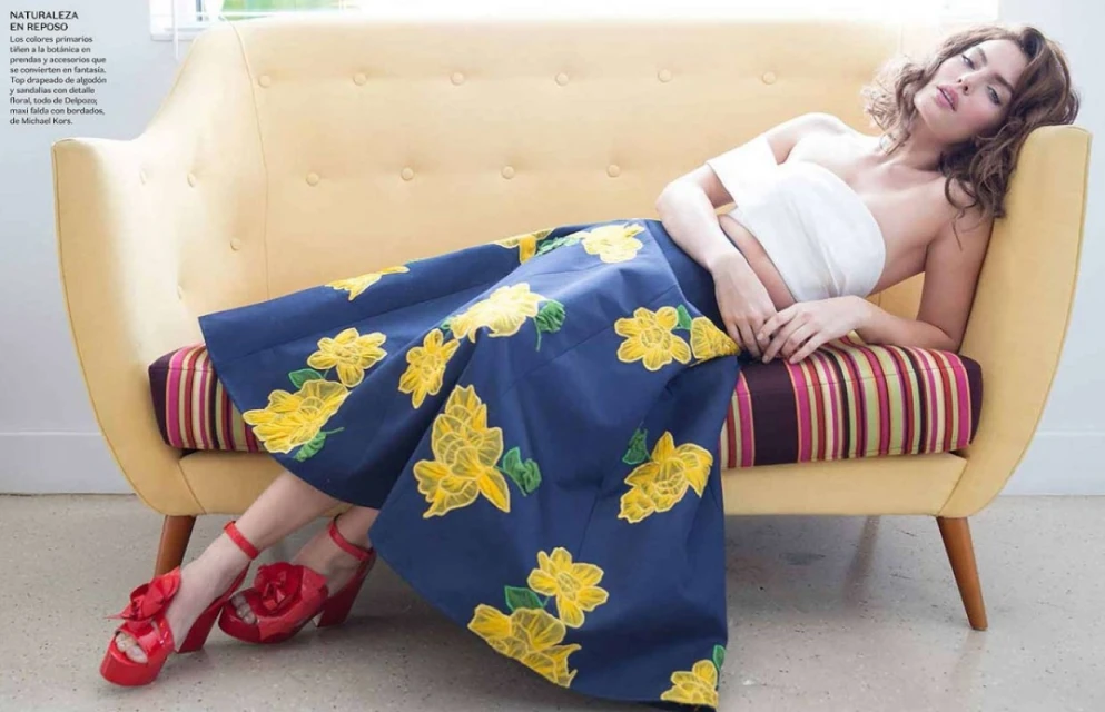 Woman in floral skirt and red shoes lounges on a yellow couch indoors; fashion editorial with bright, summery colors and retro vibes.