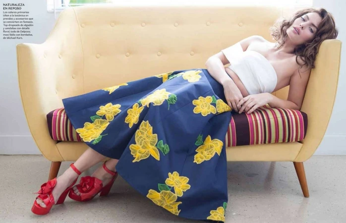 Woman in floral skirt and red shoes lounges on a yellow couch indoors; fashion editorial with bright, summery colors and retro vibes.