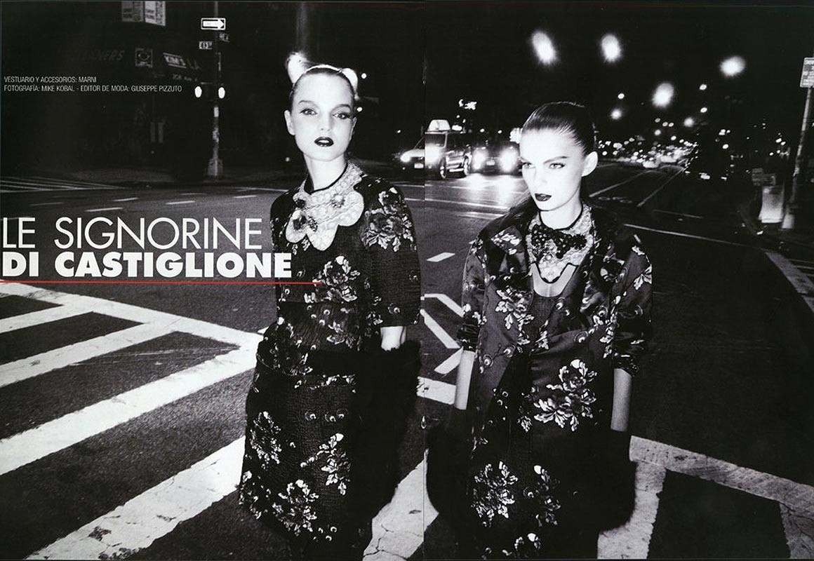Two women stand on a pedestrian crossing at night, modelling floral-patterned coats and dresses, lit by street lights and car headlights.