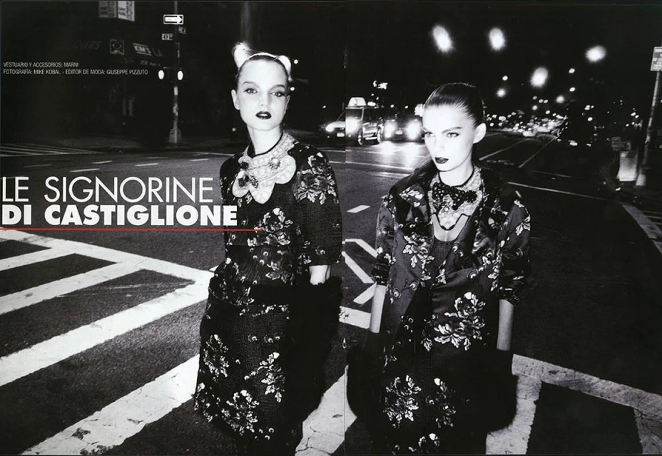 Two women stand on a pedestrian crossing at night, modelling floral-patterned coats and dresses, lit by street lights and car headlights.