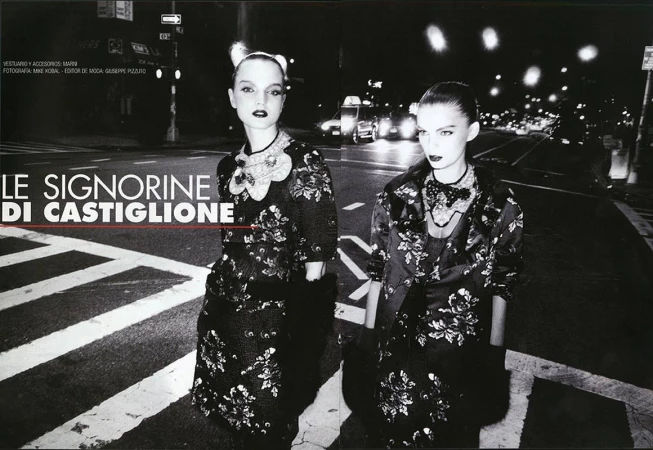 Two women stand on a pedestrian crossing at night, modelling floral-patterned coats and dresses, lit by street lights and car headlights.