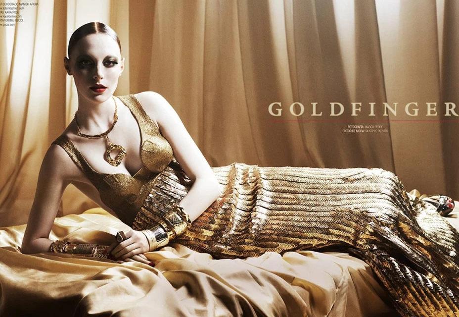 Elegant adult woman in a gold dress and jewelry lies on a gold satin surface, photographed in a studio with dramatic lighting.