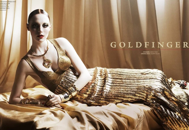 Woman in a shimmering gold dress reclining on golden draped fabric with heavy metallic jewellery and dark eye makeup.