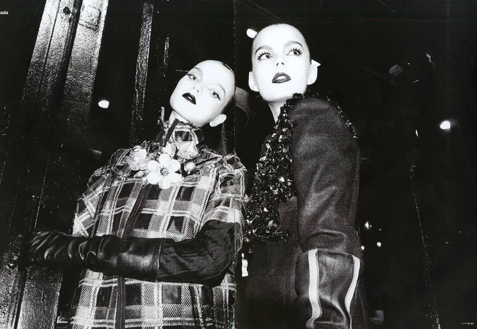 Two pale-skinned women pose indoors in avant-garde fashion with dark lipstick, plaid and floral accents, in a high-contrast black and white photograph.