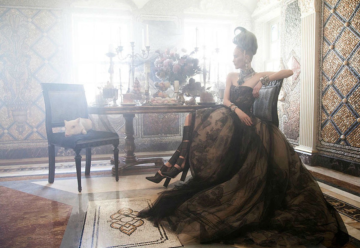 Elegant woman in ornate gown sitting in a mosaic-tiled room with a small pig on a chair, flowers, candelabras, and decorative objects.
