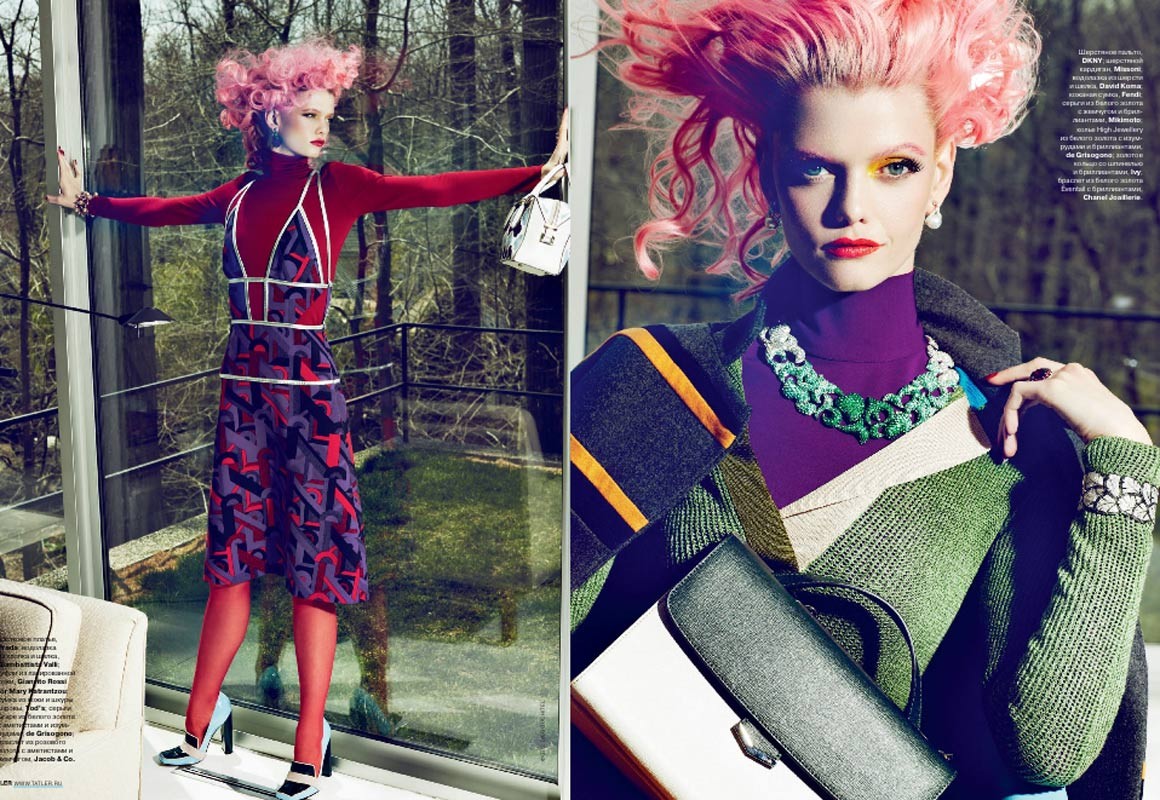 A fashion editorial shot featuring a model with pink hair wearing a purple dress and red turtleneck in front of a large window.
