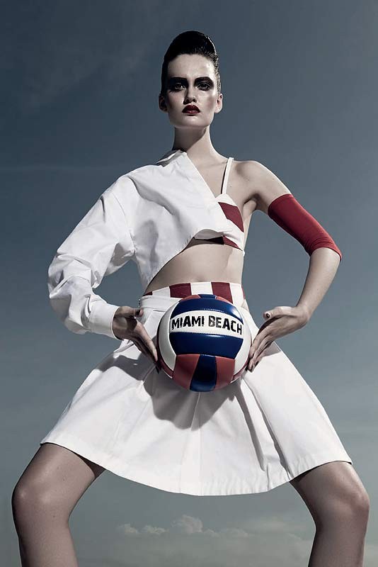 Woman strikes stylized pose outdoors in editorial fashion shot wearing white outfit, holding 'Miami Beach' volleyball against cloudy sky.