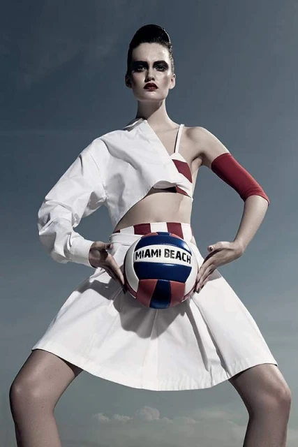Woman strikes stylized pose outdoors in editorial fashion shot wearing white outfit, holding 'Miami Beach' volleyball against cloudy sky.