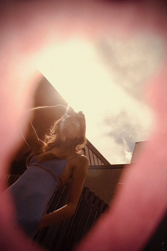Woman in a blue dress posing outside with architectural elements and a cloudy sky visible through a pink, out-of-focus foreground element.