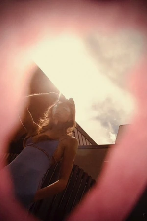 Woman in a blue dress posing outside with architectural elements and a cloudy sky visible through a pink, out-of-focus foreground element.