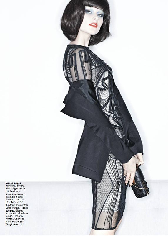 A stylish adult woman poses in a sheer black dress with intricate patterns against a bright white backdrop, capturing a high-fashion look.