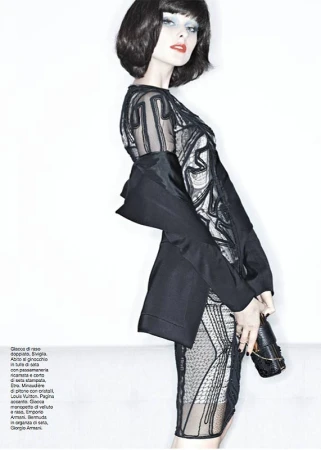 A stylish adult woman poses in a sheer black dress with intricate patterns against a bright white backdrop, capturing a high-fashion look.
