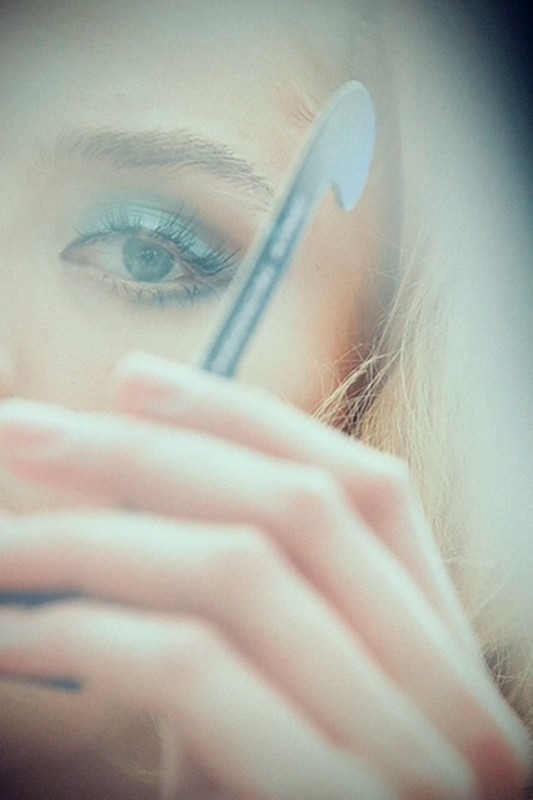 Close-up of a woman's face with bright blue eyeshadow, holding a beauty tool near her eye in a soft, dreamy aesthetic.