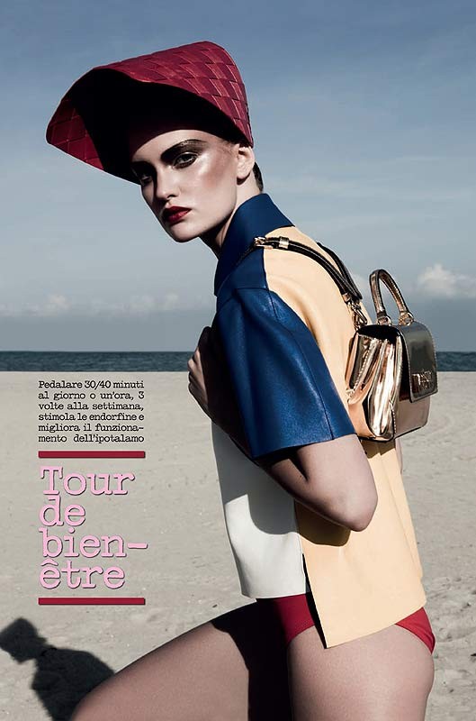Fashion editorial shot of a model on a beach wearing a colorful shirt, red bottoms, red hat, and carrying a gold handbag.