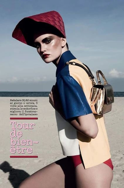 Fashion editorial shot of a model on a beach wearing a colorful shirt, red bottoms, red hat, and carrying a gold handbag.