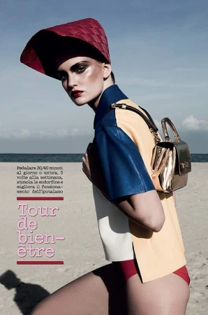 Fashion editorial shot of a model on a beach wearing a colorful shirt, red bottoms, red hat, and carrying a gold handbag.