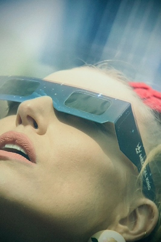 Close-up of an adult woman looking up while wearing blue, decorative eclipse-viewing glasses, bright pink headband, and round earrings.