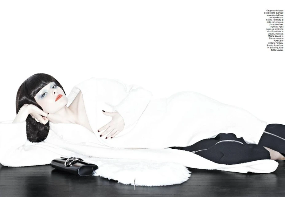 A woman with dark hair in a white robe poses on a black surface with a white rug and a black purse in a studio setting.