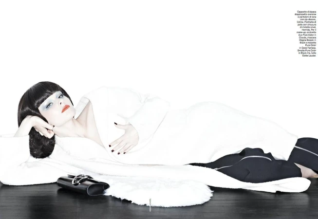 A woman with dark hair in a white robe poses on a black surface with a white rug and a black purse in a studio setting.