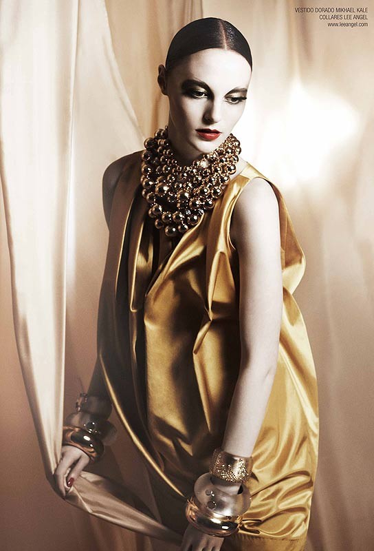 A woman with slicked-back hair poses in a gold dress with oversized, spherical jewelry. The shot uses soft, diffused lighting.