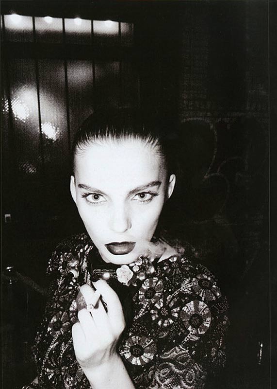 Monochrome portrait of a woman with dark lipstick, ornate jacket, holding a smoking object against a dark background. Editorial fashion.
