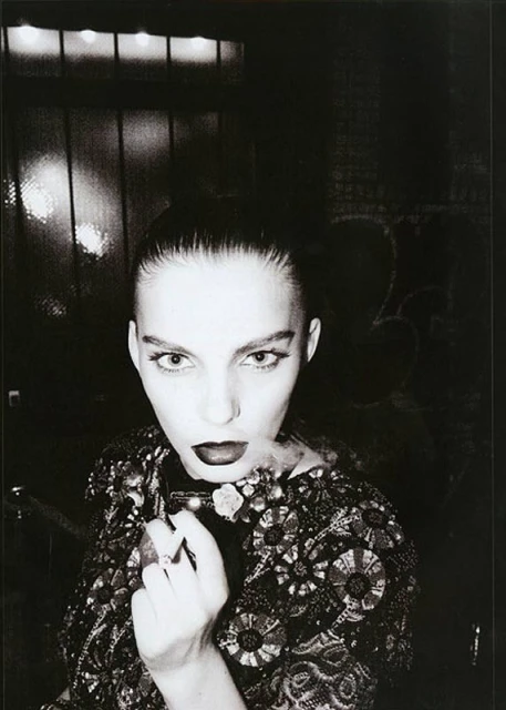 Monochrome portrait of a woman with dark lipstick, ornate jacket, holding a smoking object against a dark background. Editorial fashion.