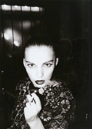 Monochrome portrait of a woman with dark lipstick, ornate jacket, holding a smoking object against a dark background. Editorial fashion.