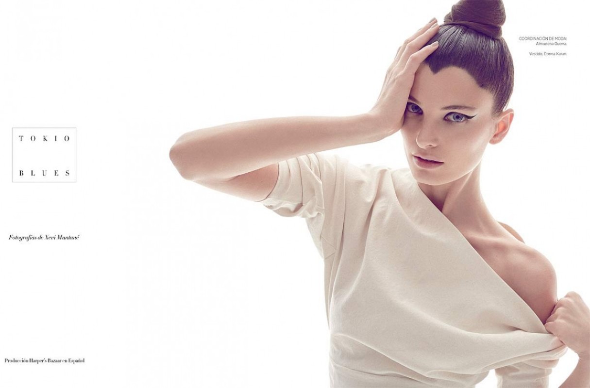 A woman in a light-colored dress poses against a white background with a stylish bun and heavy eyeliner for a fashion editorial.