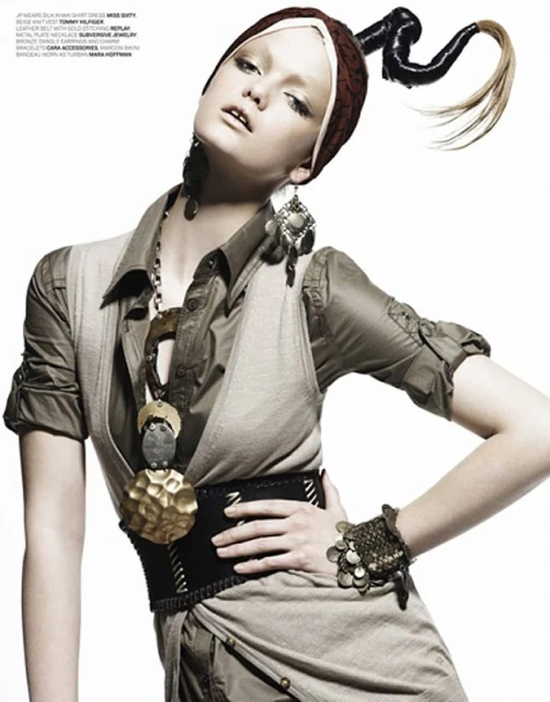 Fashion editorial shot of a female model in a brown headdress and avant-garde ponytail, statement jewelry, and a layered olive-toned dress against a white background.