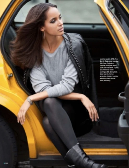 A woman with long hair in a grey sweater and black leather jacket poses in the open doorway of a yellow taxicab.