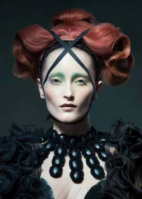 Portrait of a woman with an elaborate auburn hairstyle, black headdress, and decorative black collar, against a dark gray backdrop.
