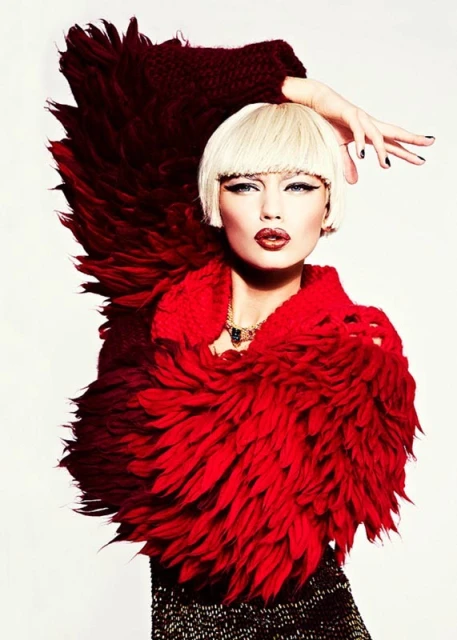 Close-up of a blond woman with heavy makeup wearing a large, feathery red and burgundy collar and dark textured shirt against white.