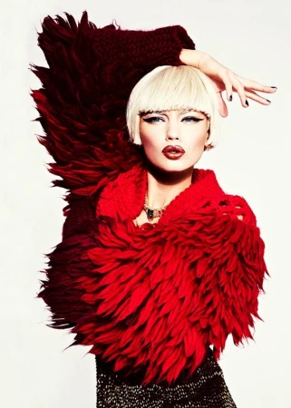 Close-up of a blond woman with heavy makeup wearing a large, feathery red and burgundy collar and dark textured shirt against white.