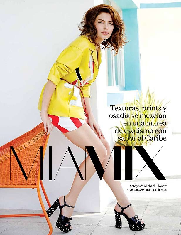 A woman in a yellow jacket and striped shorts poses next to a wicker chair with heels, in a fashion editorial image.