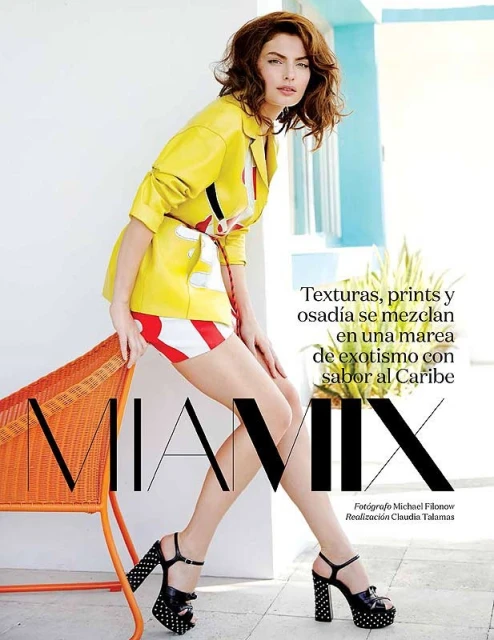 A woman in a yellow jacket and striped shorts poses next to a wicker chair with heels, in a fashion editorial image.