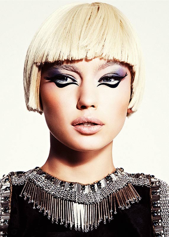 Glamorous close-up portrait of a woman with a blonde bob haircut and heavy black eye makeup, wearing a black dress with beaded trim.