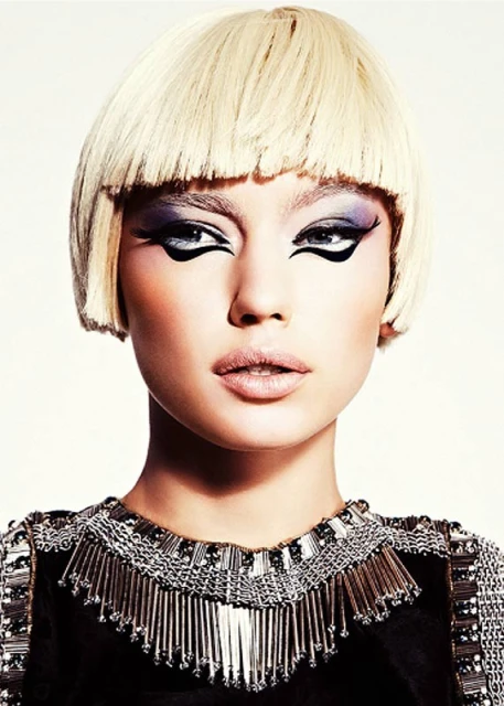 Glamorous close-up portrait of a woman with a blonde bob haircut and heavy black eye makeup, wearing a black dress with beaded trim.