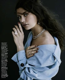 Close-up of a model wearing a light blue coat, diamond jewelry, and red nail polish against a dark background.