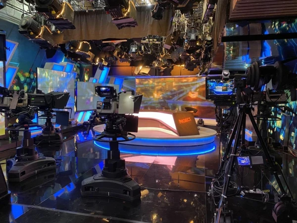 Wide shot of a modern TV studio with multiple cameras, stage lighting, video screens, and a branded news desk for 'Project'.